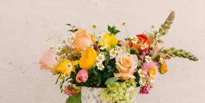 A hand holding a vase filled with a colorful bouquet of fresh flowers including roses, tulips, and daisies against a textured white wall.
