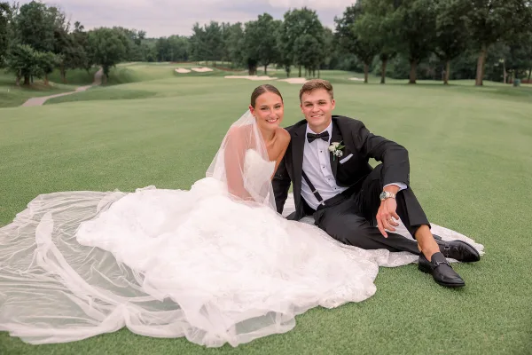 A bride and groom sitting on a blanket on a golf course lawn.