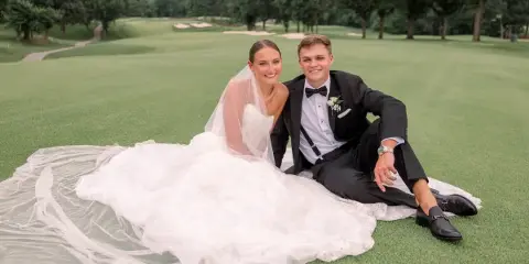 A bride and groom sitting on a blanket on a golf course lawn.
