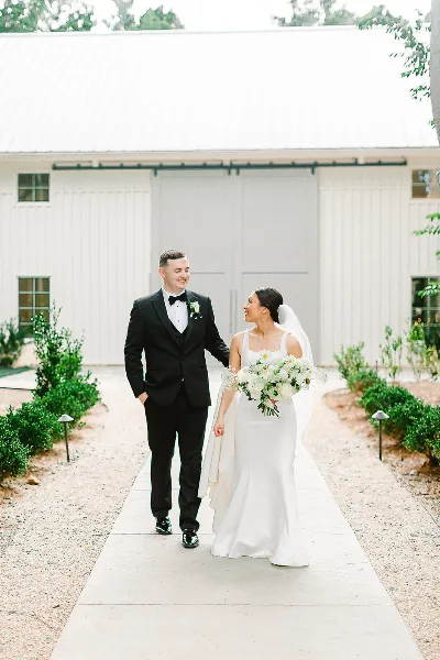 A bride and groom walking on a concrete path in front of a white barn-style wedding venue building.