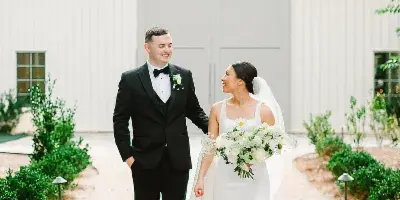 A bride and groom walking on a concrete path in front of a white barn-style wedding venue building.