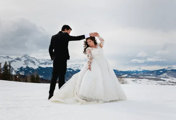 A bride and groom dancing in a snowy field with mountains in the background.