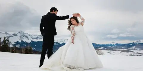 A bride and groom dancing in a snowy field with mountains in the background.