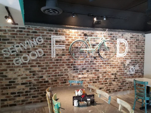Interior view of Dreez restaurant showing a dining table, chairs, and a brick wall with a bicycle decoration and 'Serving Good Food' sign.