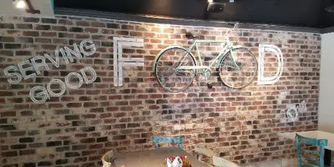 Interior view of Dreez restaurant showing a dining table, chairs, and a brick wall with a bicycle decoration and 'Serving Good Food' sign.