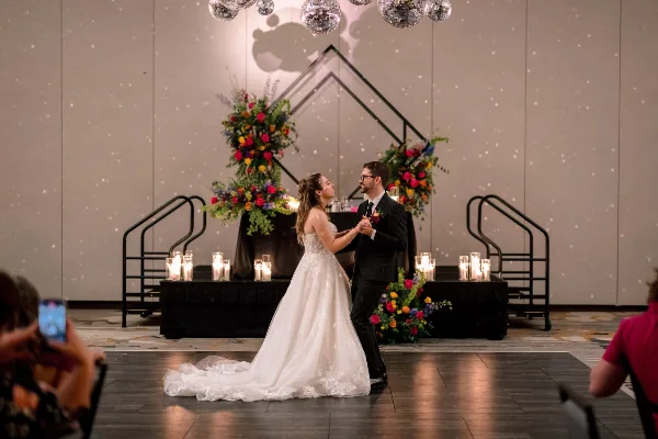 A bride and groom dancing at a wedding reception in front of a large floral arrangement and geometric metal structure.