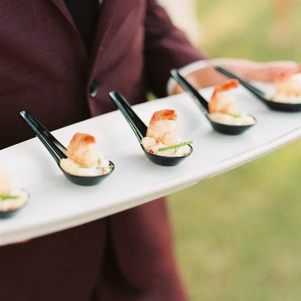 A catering server in a maroon jacket holding a white tray with individual shrimp appetizers in black spoons.