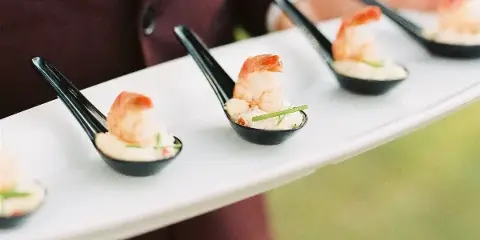 A catering server in a maroon jacket holding a white tray with individual shrimp appetizers in black spoons.