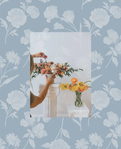 Florist arranging a colorful floral centerpiece on a white pedestal with a vase of yellow and orange flowers nearby.