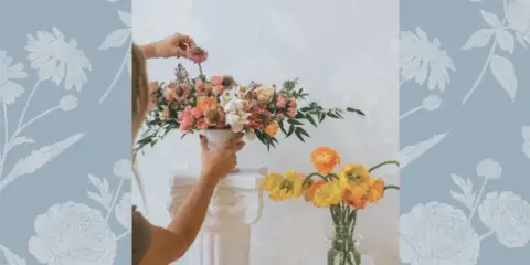 Florist arranging a colorful floral centerpiece on a white pedestal with a vase of yellow and orange flowers nearby.