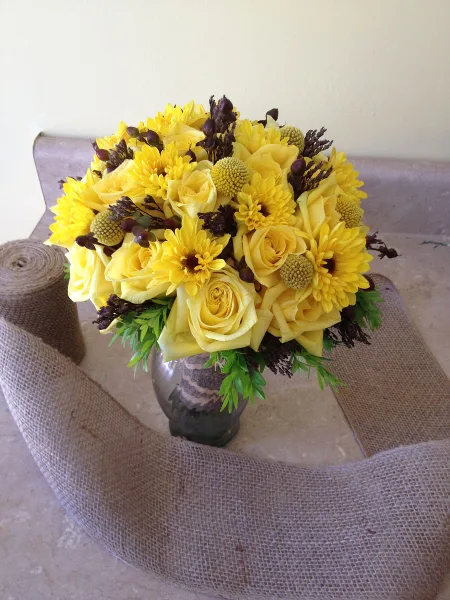 A round floral arrangement of yellow roses and chrysanthemums in a glass vase, set against a burlap backdrop.