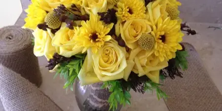 A round floral arrangement of yellow roses and chrysanthemums in a glass vase, set against a burlap backdrop.