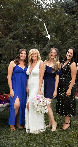 A bride in a white lace dress holding a bouquet of pink and white flowers, standing with three bridesmaids outdoors.