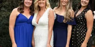 A bride in a white lace dress holding a bouquet of pink and white flowers, standing with three bridesmaids outdoors.