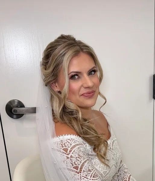 A bride with a styled updo and full makeup, wearing a lace wedding dress and veil.