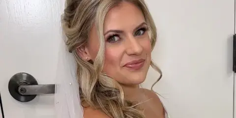 A bride with a styled updo and full makeup, wearing a lace wedding dress and veil.