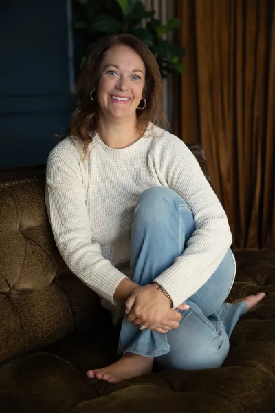 Portrait of a smiling woman with brown hair wearing a white sweater and jeans, sitting on a brown sofa.