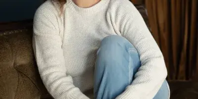Portrait of a smiling woman with brown hair wearing a white sweater and jeans, sitting on a brown sofa.