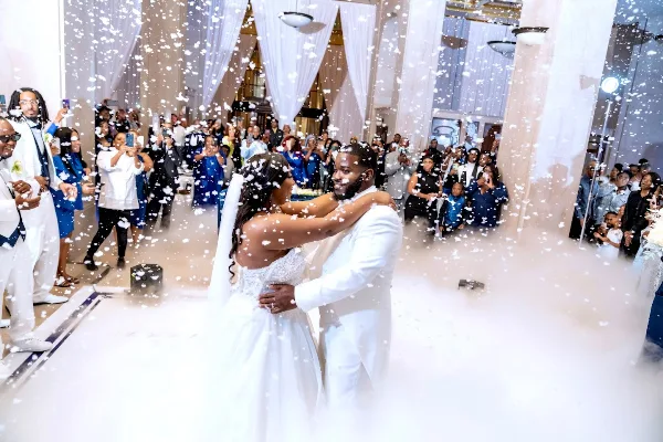 A bride and groom dancing in a grand historic venue with marble floors and high ceilings, surrounded by guests and falling confetti.
