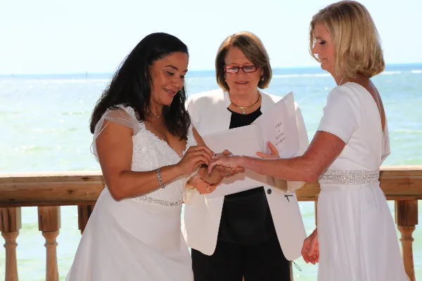 A wedding officiant in a white jacket performs a ceremony on a beach, placing a ring on a bride's hand while another woman in white looks on.