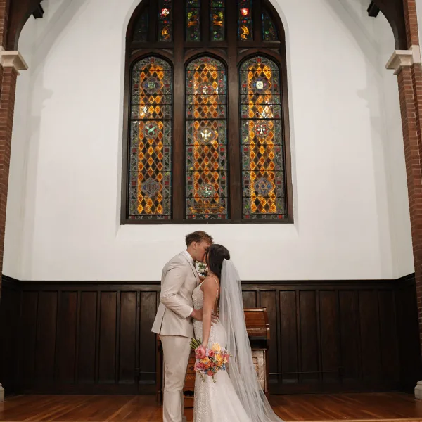 A bride and groom kissing inside a chapel with a large stained glass window.