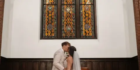 A bride and groom kissing inside a chapel with a large stained glass window.