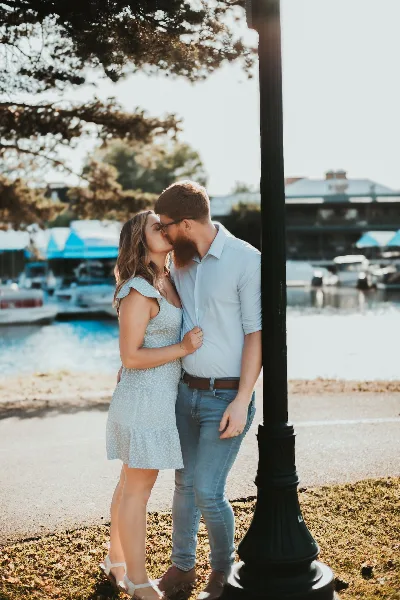 A couple sharing a kiss outdoors near a waterfront with boats in the background.