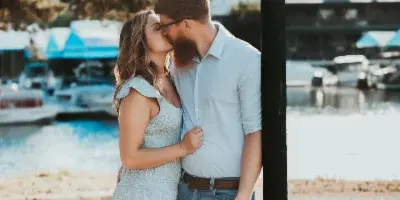 A couple sharing a kiss outdoors near a waterfront with boats in the background.