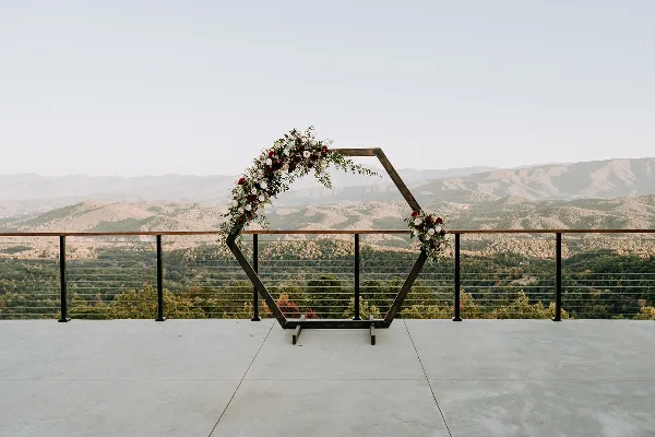 A wooden wedding arch decorated with flowers and greenery on a concrete terrace overlooking a mountain landscape.