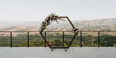 A wooden wedding arch decorated with flowers and greenery on a concrete terrace overlooking a mountain landscape.