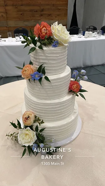A three-tiered white wedding cake decorated with fresh orange, white, and blue flowers on a table in a banquet hall.