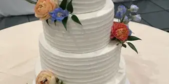 A three-tiered white wedding cake decorated with fresh orange, white, and blue flowers on a table in a banquet hall.