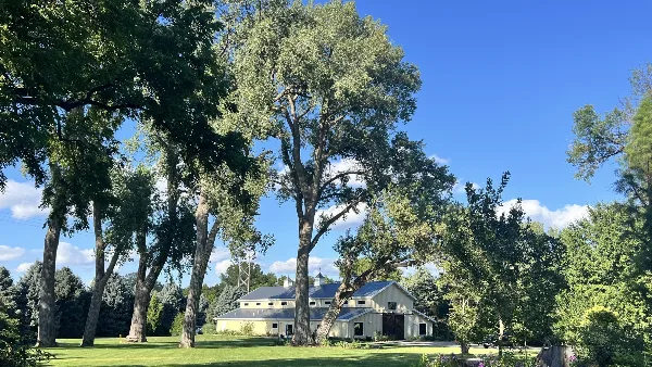 A wide shot of a large, light-colored building with a grey roof, situated on a grassy lawn surrounded by mature trees under a blue sky.