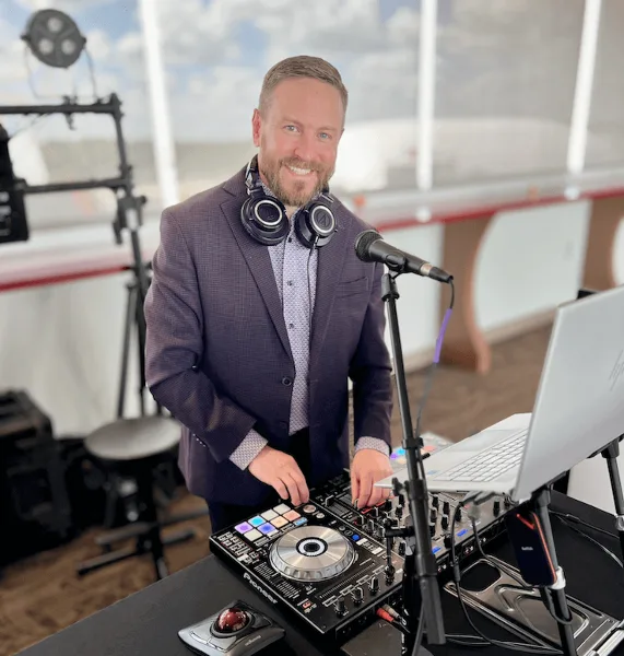 A professional DJ wearing headphones stands behind a Pioneer DJ mixer and laptop at an event venue.