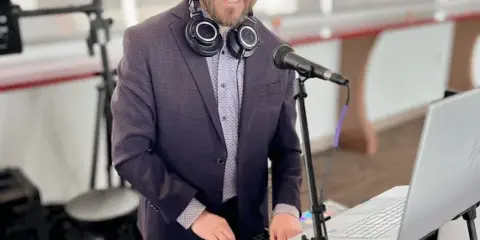 A professional DJ wearing headphones stands behind a Pioneer DJ mixer and laptop at an event venue.