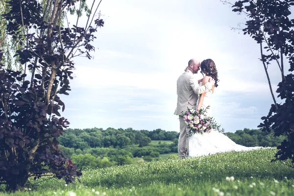 A bride and groom embracing on a grassy hill with a backdrop of green trees and a bright sky.