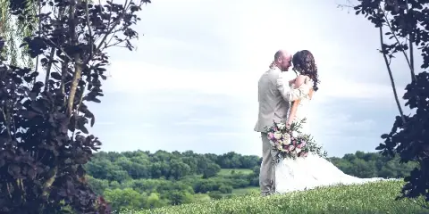 A bride and groom embracing on a grassy hill with a backdrop of green trees and a bright sky.