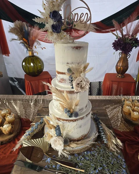 A multi-tier white wedding cake decorated with dried florals and a name topper, displayed on a table with cupcakes and floral arrangements.
