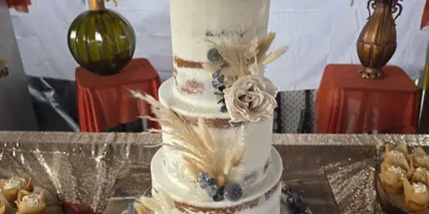 A multi-tier white wedding cake decorated with dried florals and a name topper, displayed on a table with cupcakes and floral arrangements.
