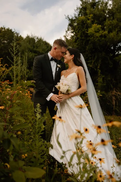 A bride and groom standing together in a field with wildflowers, the groom in a black tuxedo and the bride in a white wedding dress holding a bouquet.