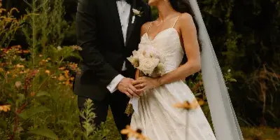 A bride and groom standing together in a field with wildflowers, the groom in a black tuxedo and the bride in a white wedding dress holding a bouquet.