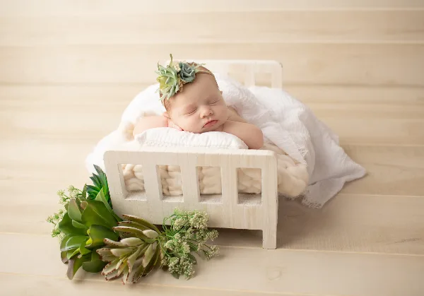 A sleeping newborn baby wearing a succulent headband, lying in a small white wooden bed with greenery props on a light wood floor.