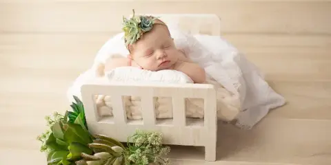 A sleeping newborn baby wearing a succulent headband, lying in a small white wooden bed with greenery props on a light wood floor.