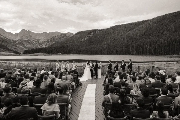 A black and white photograph of a wedding ceremony on a wooden deck overlooking a mountain lake, with the bride and groom at the altar and guests seated in rows.