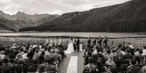 A black and white photograph of a wedding ceremony on a wooden deck overlooking a mountain lake, with the bride and groom at the altar and guests seated in rows.