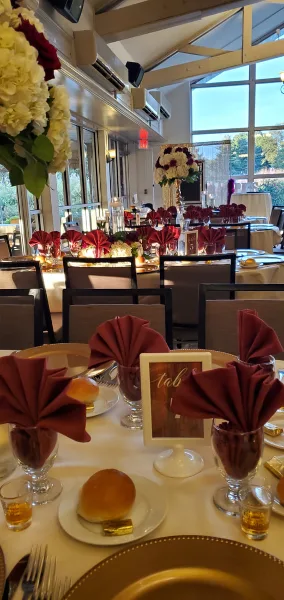 Catering table set with bread rolls, folded burgundy napkins, glassware, and floral centerpieces in a bright event venue.