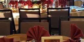 Catering table set with bread rolls, folded burgundy napkins, glassware, and floral centerpieces in a bright event venue.