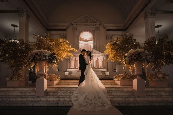 A bride and groom standing together in a grand, ornate hall with large floral arrangements.