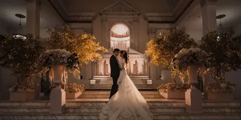 A bride and groom standing together in a grand, ornate hall with large floral arrangements.