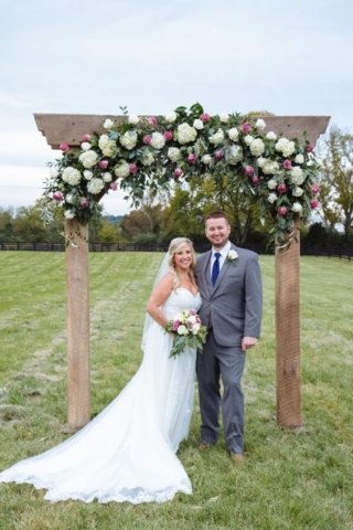 A bride and groom standing under a floral arch in a grassy field.